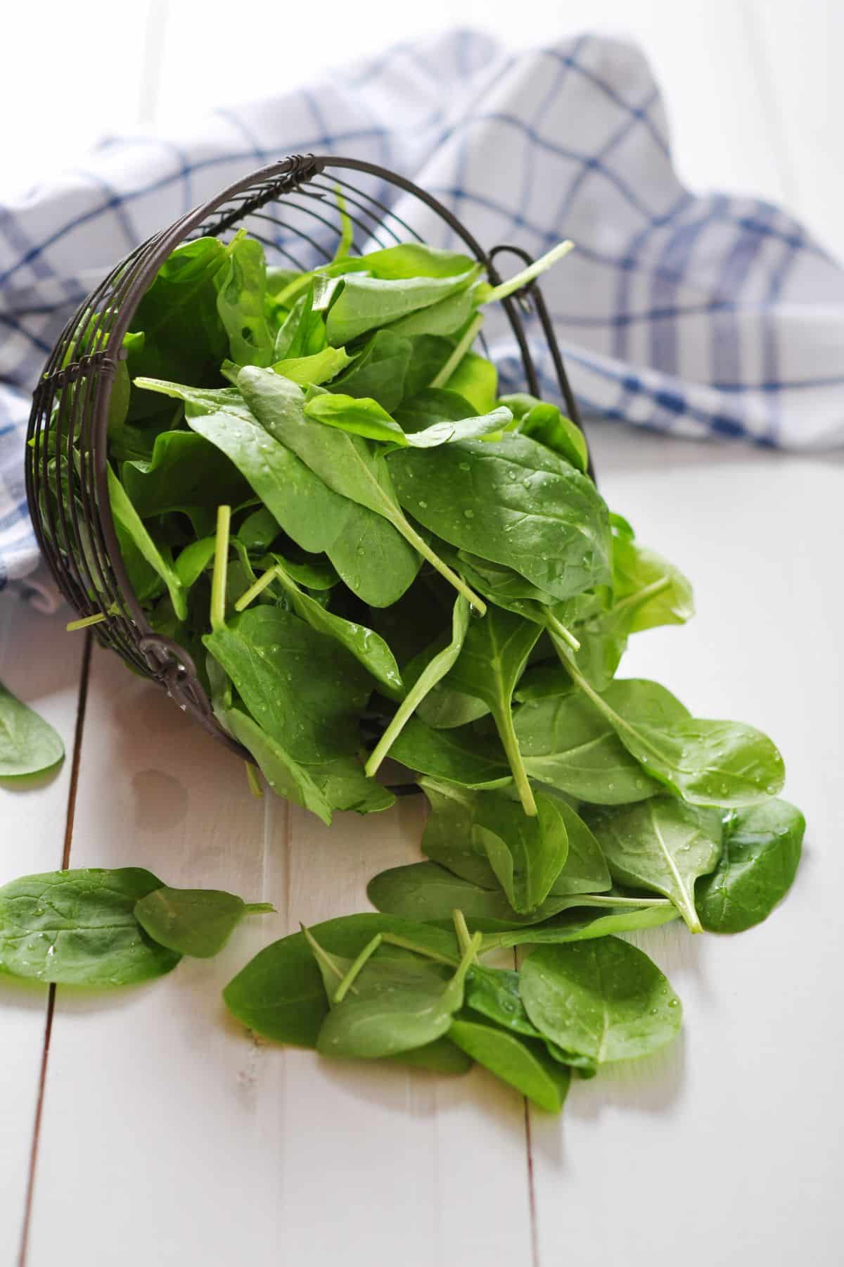 Basket of baby spinach leaves spilling onto wooden surface.