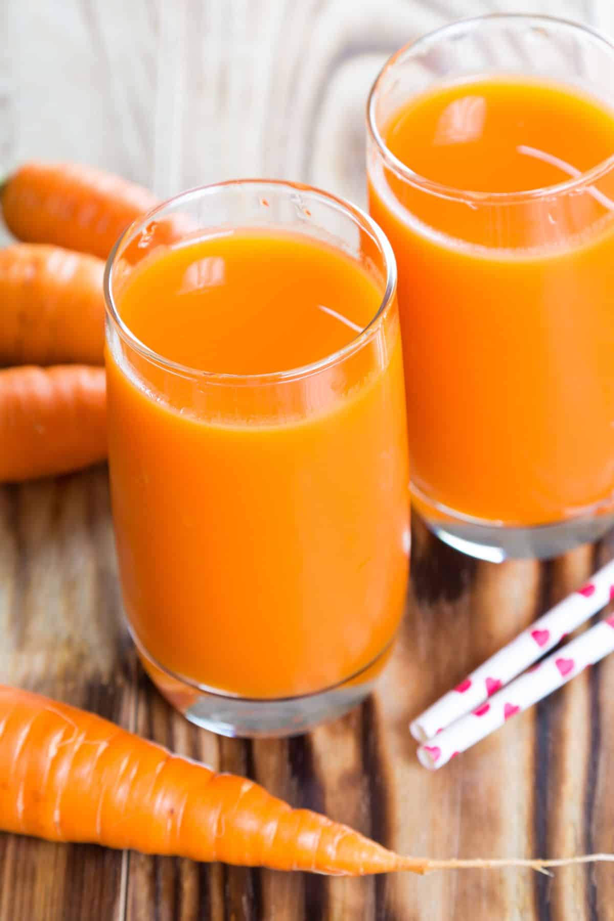 two glasses of carrot juice surrounded by whole carrots on a table.