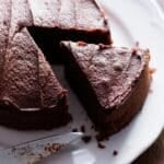 A round dairy-free chocolate cake on a white plate with a slice cut.