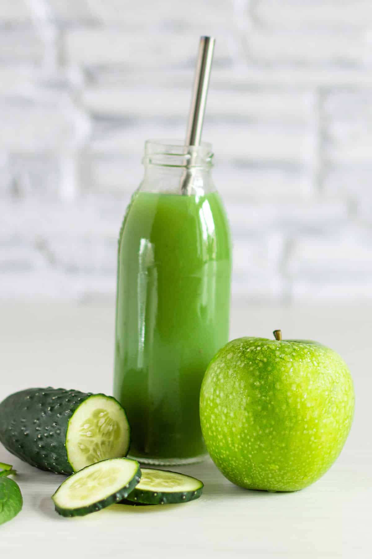 A tall thin milk bottle of green juice next to an apple and cucumber.