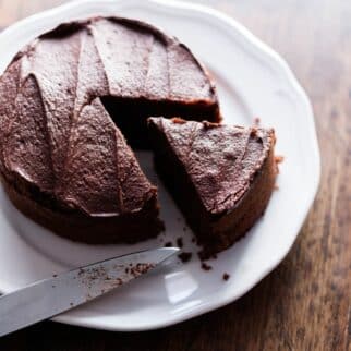 A round dairy-free chocolate cake on a white plate with a slice cut.