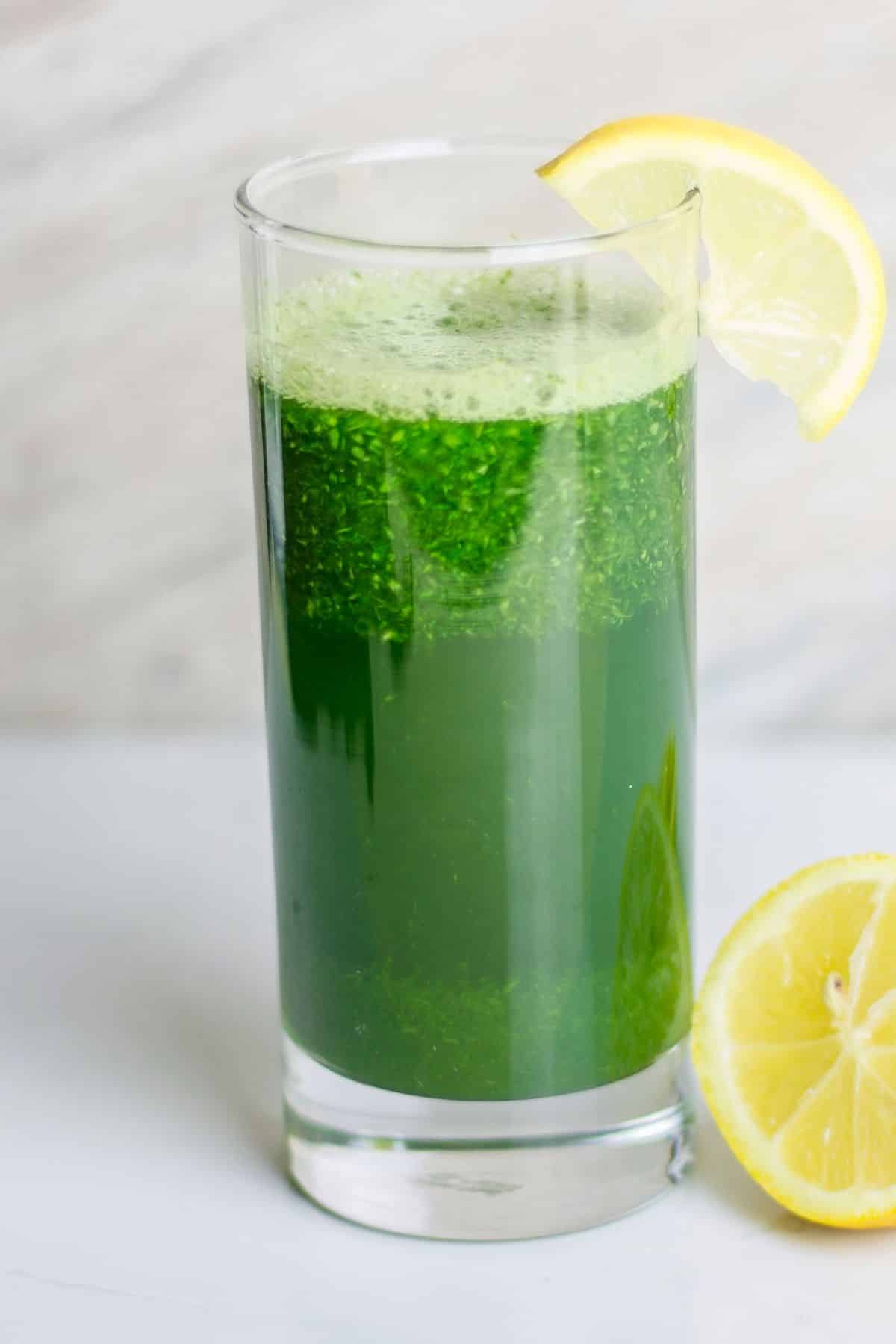 A skinny glass of spinach juice with a lemon wedge on the rim.