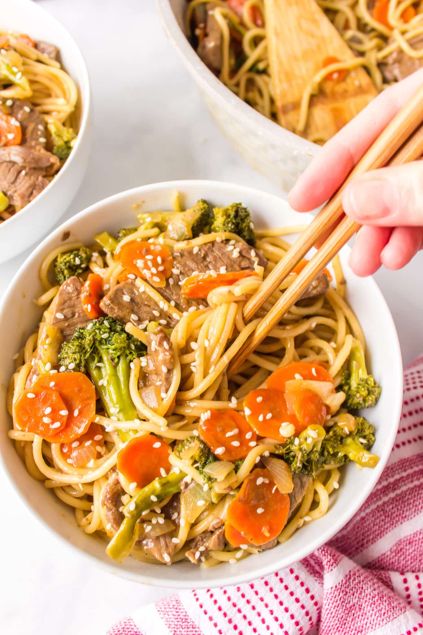 A hand holding a pair of chopsticks over a bowl of teriyaki beef noodles.