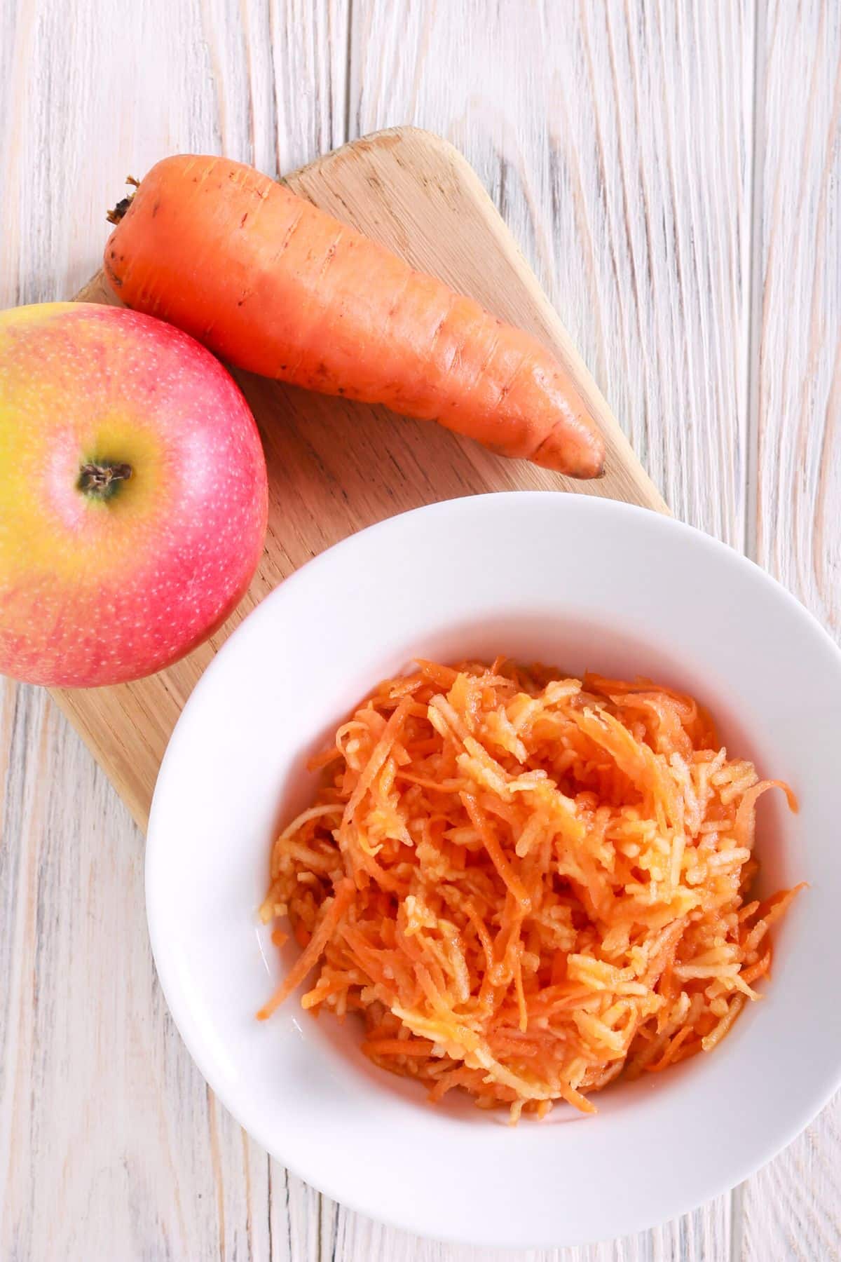Grated carrot in a bowl next to an apple and a carrot.