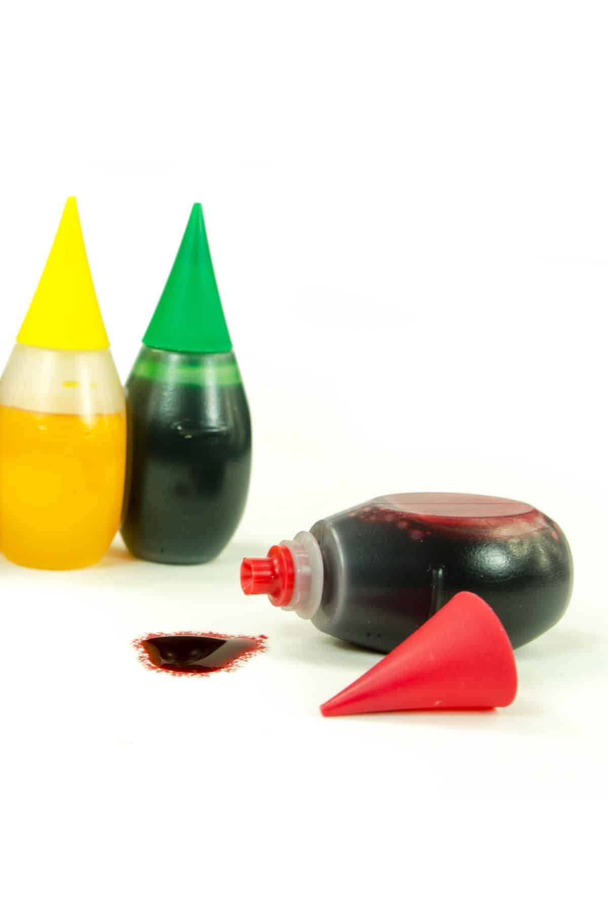 yellow, green and red food coloring bottles, with the red turned on its side spilling out.