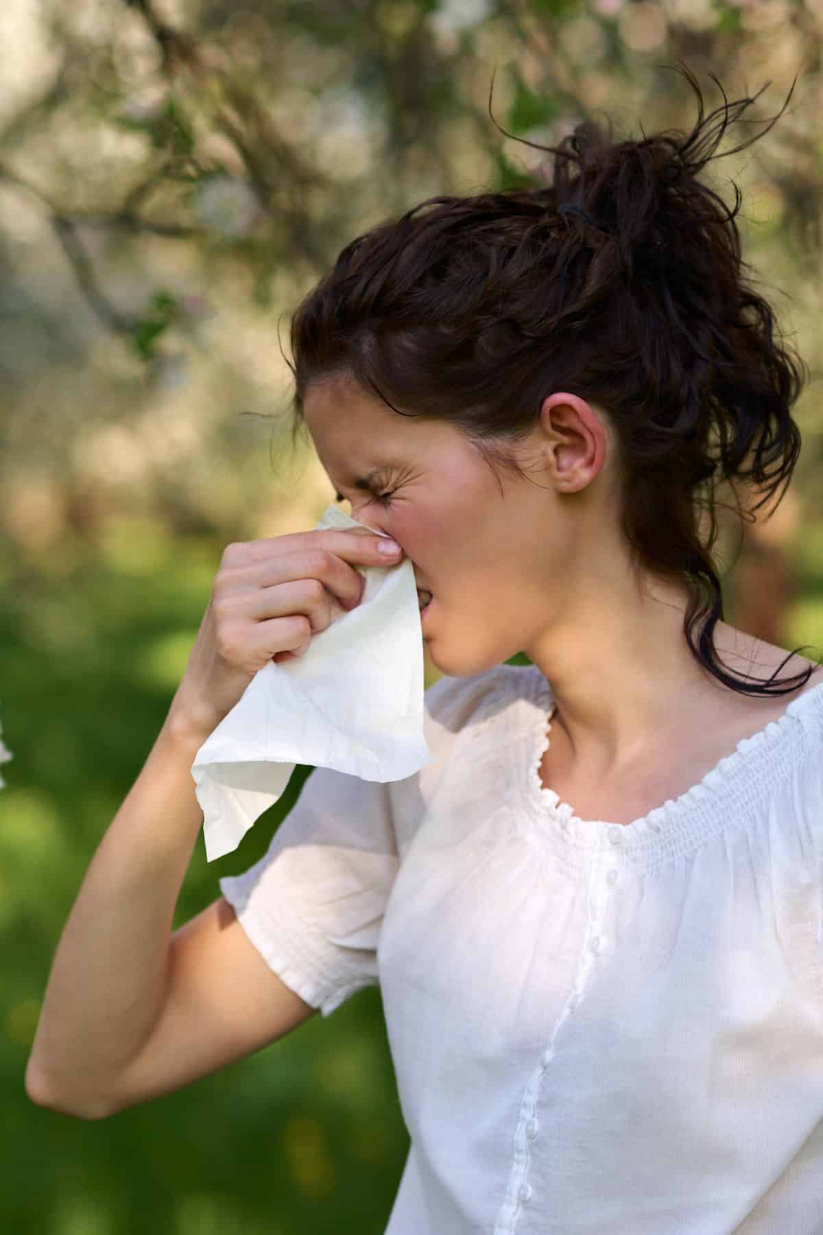 a woman outside holding her nose with a tissue and squinting her eyes.