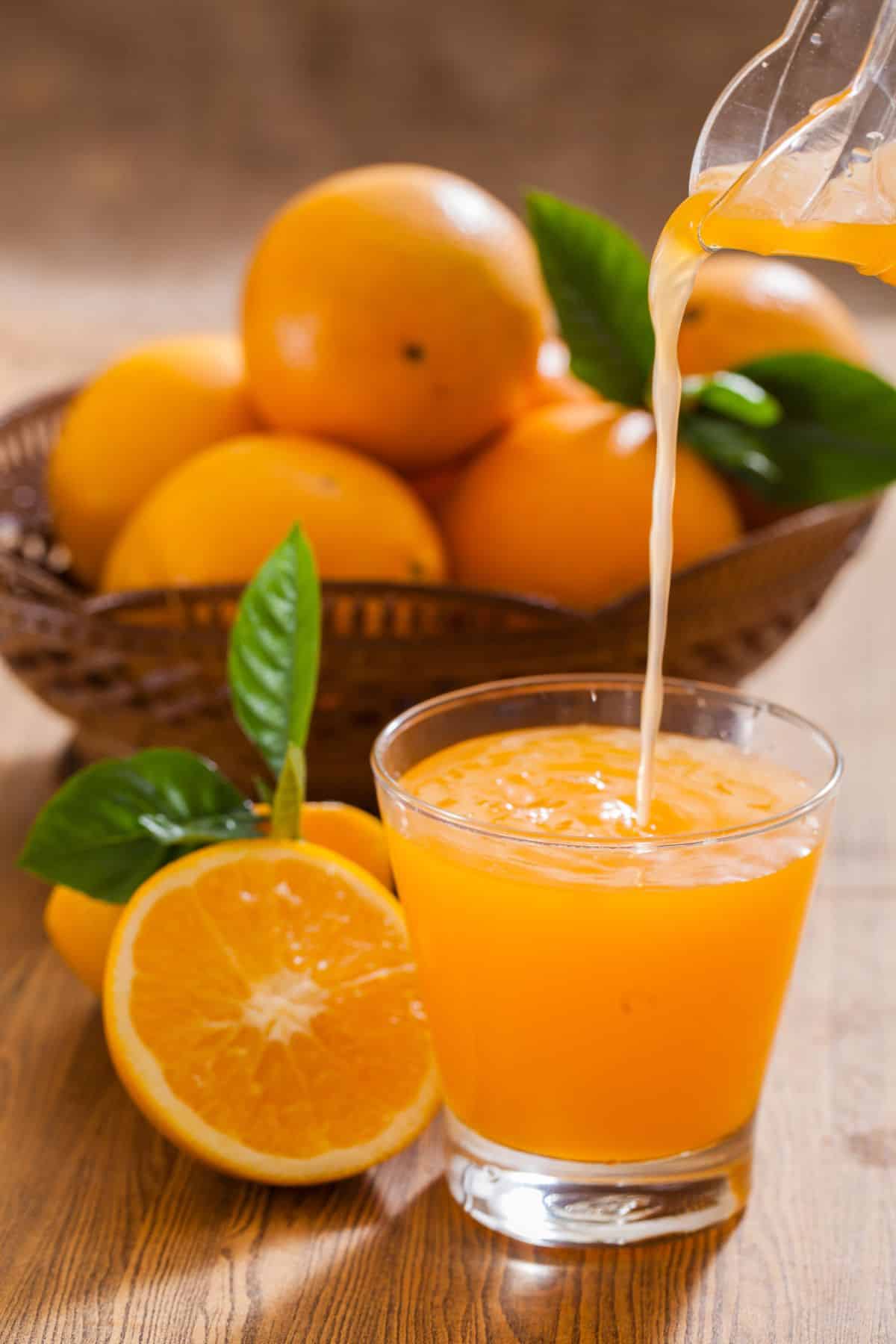 a glass of orange juice being poured in front of a bowl of oranges.