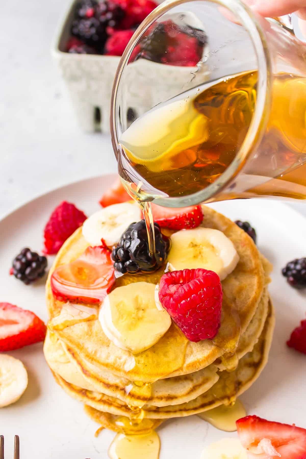 syrup being poured over a stack of pancakes topped with fruit.
