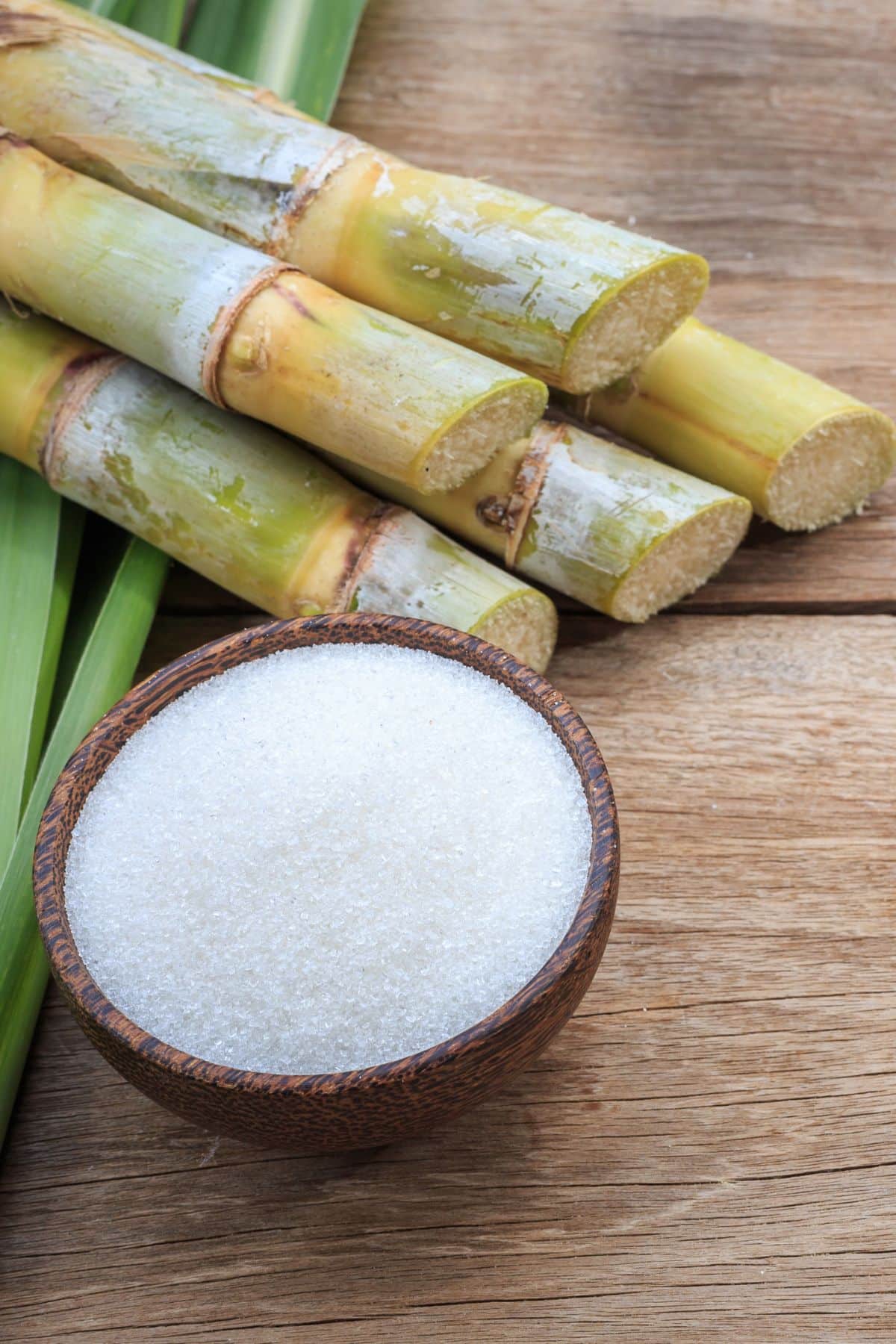 a wooden bowl of cane sugar next to sugar cane.